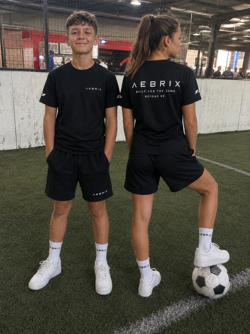 Two children modeling black Aebrix athletic t-shirts and shorts on an indoor artificial turf field, with one child showing the back of the shirt, which reads 'AEBRIX