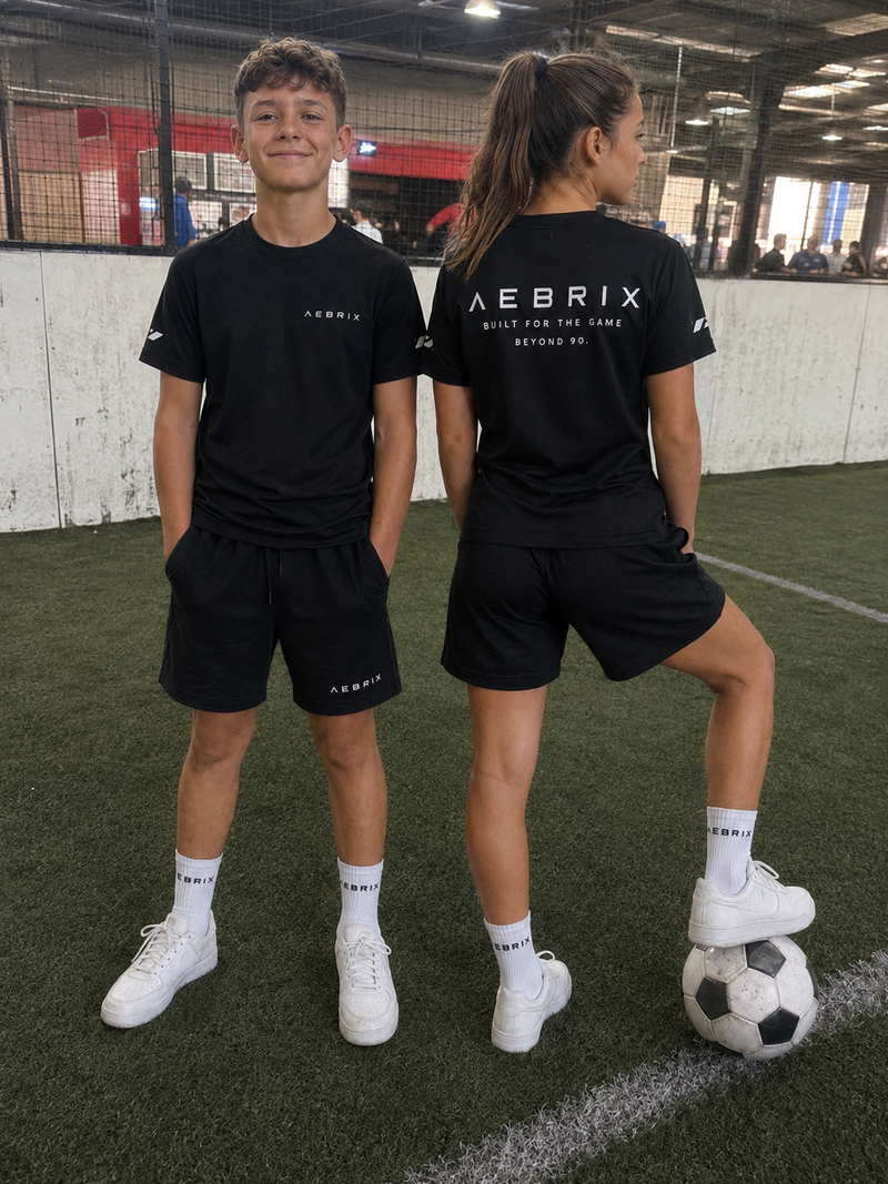 Two children modeling black Aebrix athletic t-shirts and shorts on an indoor artificial turf field, with one child showing the back of the shirt, which reads 'AEBRIX